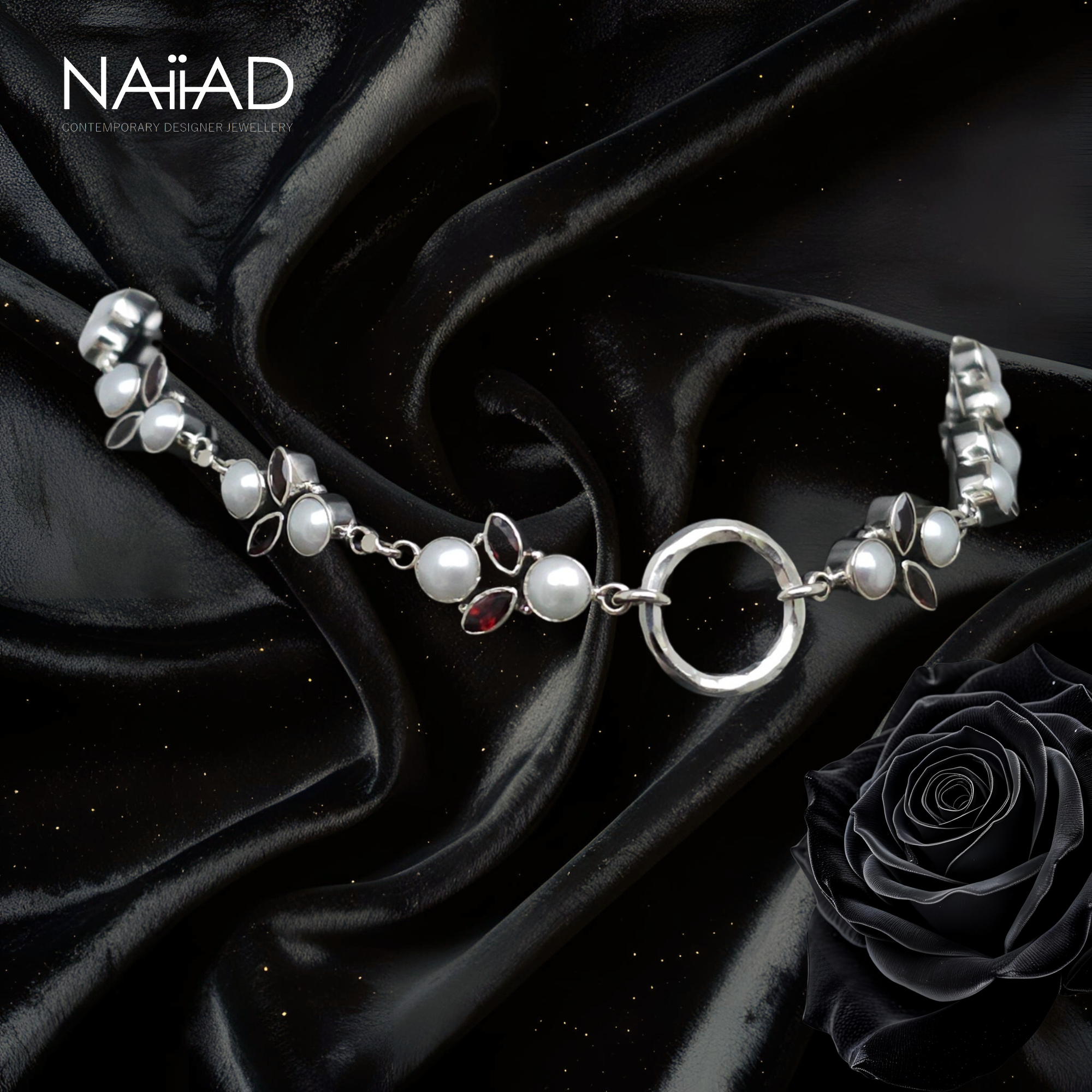 Silver necklace with pearls and a red gemstone on black satin fabric, featuring the brand 'Naiiad'.